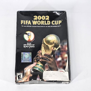 May include: A black rectangular video game box for 2002 FIFA World Cup. The cover features gold text, the official tournament logo, a holographic sticker, and an image of hands holding the gold trophy. An ESRB Everyone E rating is in the corner.