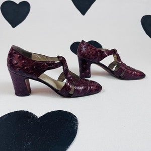 May include: A pair of burgundy leather heeled shoes with a t-strap design. The shoes have a textured surface and a rounded toe. The heels are approximately 3 inches tall. The background is white with black heart shapes.