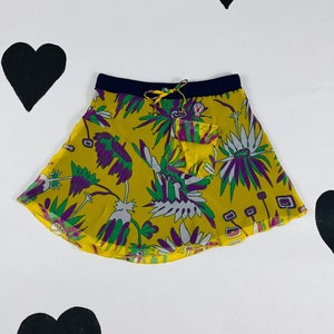 May include: A yellow skirt with a floral pattern in green, purple, and white. The skirt has a navy blue waistband and a small pocket. The background is white with black heart shapes.