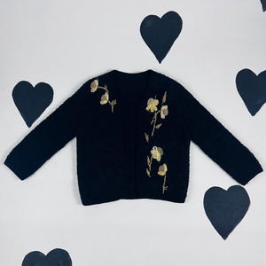 以下が含まれることがあります： 刺繍された花のディテールが施された黒いカーディガン。カーディガンはシンプルなデザインで、柔らかくふわふわした素材で作られています。