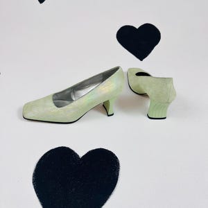 May include: A pair of light green high-heeled shoes with a square toe. The shoes have a shimmery finish and are on a white background with black hearts.