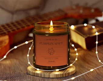 May include: A lit candle in a glass jar with a label that reads "Pumpkin Spice". The label also features a maple leaf illustration and the words "Cinnamon & Vanilla" and "Handmade". The candle sits on a wooden coaster, with string lights in the background.