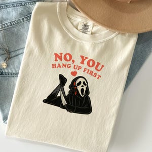 以下が含まれることがあります： 「NO, YOU HANG UP FIRST」の赤い文字が入ったクリーム色のTシャツ。黒いローブとマスクを着けたキャラクターが電話を持っている漫画が描かれています。赤いハートが単語の間にあります。このシャツは、ブルージーンズと茶色の帽子と組み合わせています。