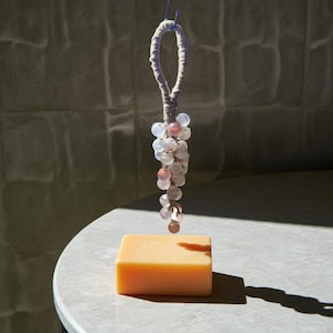May include: A rectangular, light orange bar of soap sits on a white surface. Above it hangs a decorative beaded ornament with white and pink beads, suspended by a loop of white fabric and blue string. The soap's shadow is cast on the surface.