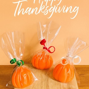 Puede incluir: Tres jabones naranjas con forma de calabaza, envueltos en bolsas de celofán y atados con cintas verdes, rojas y blancas. Están sobre una tabla de madera, con "Happy Thanksgiving" escrito encima.
