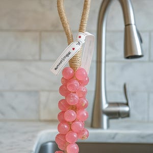 Può includere: Una spugna rosa piena di sapone a forma di grappolo d'uva, appesa a un anello di corda naturale. Un nastro bianco con testo e un piccolo righello sono attaccati. Lo sfondo comprende un rubinetto in acciaio inossidabile e una parete piastrellata bianca.