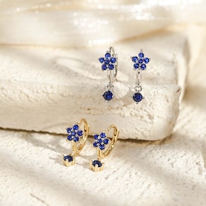 Op de afbeelding: Twee paar kleine oorringen, een zilverkleurige en een goudkleurige. Elke oorbel heeft een bloemvormige bedel bezet met kleine blauwe steentjes en een enkele bungelende blauwe steen. De oorbellen worden gepresenteerd als twee afzonderlijke sets.