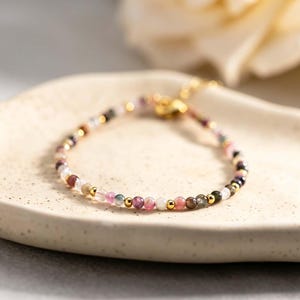 Op de afbeelding: Een delicate armband met kleine, veelkleurige toermalijnkralen afgewisseld met piepkleine, gepolijste goudkleurige metalen bolletjes. De armband is voorzien van een goudkleurige metalen sluiting en ligt in een cirkelvorm op een textuurrijk, gebroken wit oppervlak.