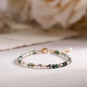 以下が含まれることがあります： 小さな緑、白、金色のビーズが特徴の繊細なビーズブレスレット。 ブレスレットには金の留め金と小さな真珠のチャームが付いています。 ジュエリーは、背景にソフトフォーカスのバラがある白い表面に表示されています。