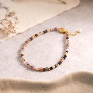 Puede incluir: Una pulsera delicada que presenta pequeñas cuentas de piedras preciosas multicolores intercaladas con diminutas esferas de metal dorado. La pieza de joyería incluye un cierre de mosquetón dorado y una cadena extensora corta con una cuenta.
