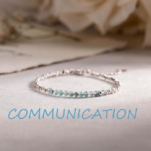 Op de afbeelding: Een delicate armband met een reeks kleine, gefacetteerde lichtblauwe kralen afgewisseld met gepolijste zilverkleurige metalen kralen. Het woord COMMUNICATION staat in blauwe hoofdletters onder de armband op het oppervlak gedrukt.
