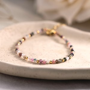 Puede incluir: Delicada pulsera de cuentas con una mezcla de piedras preciosas de colores, incluyendo rosa, morado y verde, intercaladas con pequeñas cuentas doradas. La pulsera se exhibe sobre un plato de cerámica claro y texturizado, mostrando su intrincado diseño.