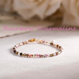Puede incluir: Delicada pulsera con pequeñas cuentas multicolores en tonos rosa, verde y dorado. La pulsera está ensartada en una fina cadena dorada. El texto "Rainbow Tourmaline" está escrito en rosa en cursiva. Se muestra sobre una superficie clara.
