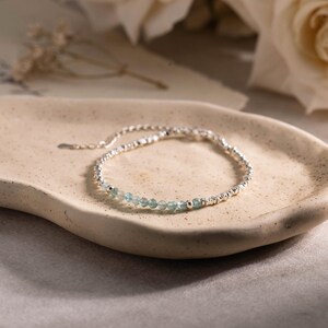 Op de afbeelding: Een verfijnde armband bestaande uit een reeks kleine, gefacetteerde lichtblauwe edelsteenkralen afgewisseld met piepkleine, gepolijste zilverkleurige metalen bolletjes. De armband is voorzien van een dunne zilverkleurige kettingverlenging.