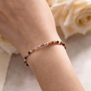 May include: Delicate beaded bracelet featuring small, colorful gemstones in shades of pink, green, brown, and gold. The bracelet is strung on a thin, gold-colored chain. A simple and elegant piece of jewelry.