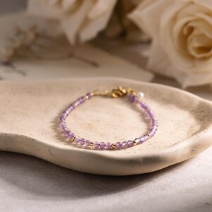 Puede incluir: Una delicada pulsera con pequeñas cuentas redondas de color púrpura claro y detalles dorados. La pulsera se exhibe sobre un plato crema de forma orgánica, con rosas blancas en el fondo.