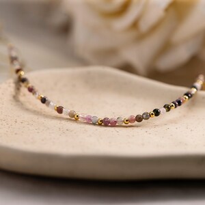 Puede incluir: Delicado collar de cuentas con pequeñas piedras preciosas de colores, en tonos rosa, morado, verde y marrón. El collar tiene detalles dorados y se muestra sobre una superficie neutra. Es una joya.