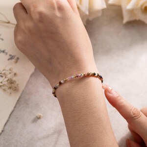 Puede incluir: Una pulsera delicada que presenta una serie de pequeñas cuentas de piedras preciosas multicolores intercaladas con diminutas cuentas espaciadoras de metal en tono dorado pulido. Las cuentas muestran tonos rosas, verdes y oscuros.