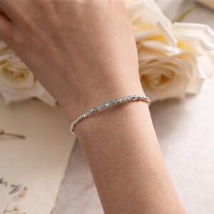 Op de afbeelding: Een verfijnde armband bestaande uit een enkele streng van kleine, gefacetteerde lichtblauwe edelsteenkralen afgewisseld met piepkleine, reflecterende zilverkleurige metalen tussenkralen voor een subtiele en elegante uitstraling.