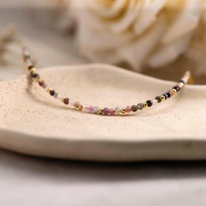 Puede incluir: Una delicada pulsera de cuentas con pequeñas piedras naturales multicolores en tonos rosa, verde y transparente, intercaladas con diminutas cuentas espaciadoras de metal dorado pulido en un patrón simétrico repetitivo.
