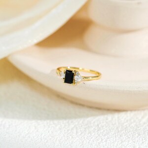 May include: A gold ring featuring a central rectangular black gemstone flanked by two smaller pear-shaped clear stones. The band is thin and polished, with the black stone held in a four-prong setting, creating a balanced and refined appearance.