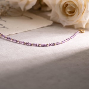 May include: Delicate amethyst bead necklace with gold-colored accents. The necklace is strung with small, round, purple beads interspersed with tiny gold beads. The clasp is also gold. The background features soft lighting and white roses.