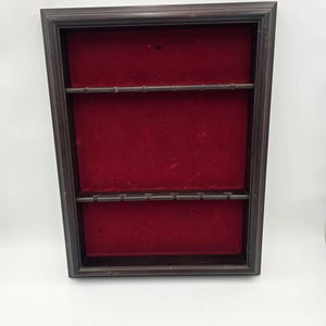 May include: A rectangular wall-mounted display case featuring a dark brown wooden frame and a deep red velvet-lined interior. The case includes two horizontal shelves, each equipped with six small notches designed for holding items securely in place.