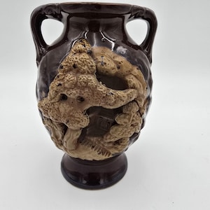 Può includere: Un vaso in ceramica marrone con due manici e un design a rilievo strutturato. Il rilievo presenta un motivo organico marrone chiaro su uno sfondo marrone più scuro. Il vaso ha un corpo arrotondato e una base a piedistallo.