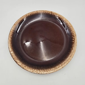 May include: A round, brown ceramic plate with a glossy finish. The plate has a darker brown center and a lighter, textured rim. The plate is likely for serving food.
