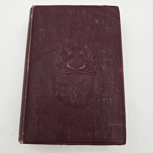 May include: A vintage, burgundy-colored book with a debossed emblem on the cover. The book has a rectangular shape and shows signs of wear, suggesting it is an antique or a well-loved item. The cover has a textured appearance.