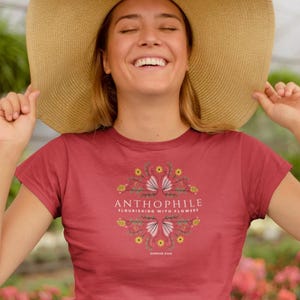 May include: A red t-shirt with the word "ANTHOPHILE" and the phrase "FLOURISHING WITH FLOWERS" printed on the front. The shirt features a floral design in yellow and pink. The person is wearing a large straw hat.