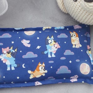 May include: A navy blue rectangular pillow with a cartoon character design, clouds, moons, and stars. The pillow features various characters in different poses. A white crocheted teddy bear and a white decorative object are also present in the image.