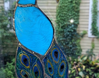 May include: A stained glass peacock suncatcher with blue, green, and gold accents. The peacock's tail feathers are spread out in a fan shape.