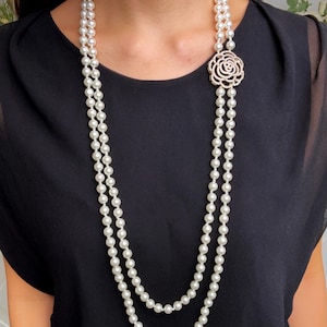 May include: A long, double-strand pearl necklace with a decorative rose-shaped pendant. The necklace features numerous white pearls and a sparkling, ornate pendant. The jewelry is displayed against a black background.