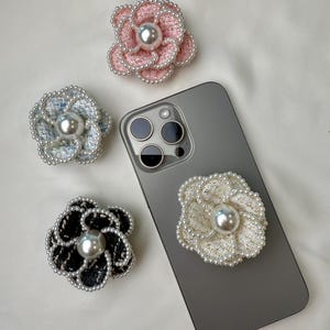 Pode incluir: Quatro suportes de telefone em forma de flor em tecido tweed rosa, azul, preto e branco, cada um com um centro de pérola e bordas com contas. Um suporte está preso a um smartphone cinza.