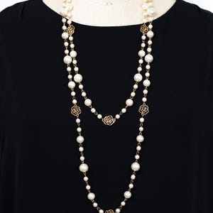 Könnte beinhalten: Eine lange, mehrreihige Perlenkette mit goldfarbenen Rosenakzenten. Die Halskette hat unterschiedlich große Kunstperlen und filigrane Kettenglieder. Die Halskette wird vor einem schwarzen Hintergrund präsentiert.