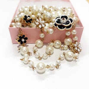 Puede incluir: Una colección de collares y pendientes de perlas con detalles dorados y adornos florales. Los pendientes y collares presentan grandes perlas redondas de color blanco roto y diseños de flores en blanco y negro. Las joyas se exhiben en una caja rosa.