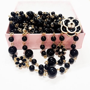 May include: Black beaded necklace with gold-tone chain and flower accents. The necklace features large, glossy black beads and decorative flower pendants with white and gold details. Presented in a pink box.