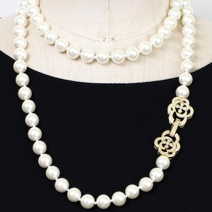 Peut inclure: Un long collier de perles blanches avec un fermoir en forme de fleur doré orné de petits cristaux transparents. Le collier est drapé sur un mannequin avec un haut noir.