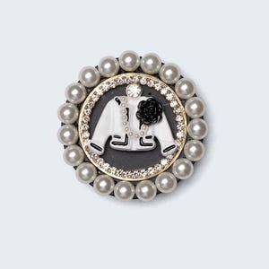 May include: A round brooch featuring a white jacket design with a black rose and pearl accents. The jacket is set against a black background, surrounded by a gold-toned frame with clear crystals and a border of faux pearls.