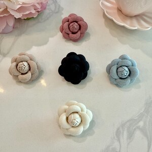 Pode incluir: Uma coleção de enfeites de flores de camélia de tecido em várias cores. As flores são em tons de rosa, bege, preto e azul claro. Cada flor tem um design de pétalas detalhado e em camadas e um centro texturizado.