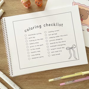 May include: A spiral-bound coloring checklist notebook with the words "coloring checklist" at the top. The checklist includes activities like "beachside cycling" and "boba break." Two small toy figures and several colored pens are also visible.