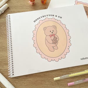 May include: A spiral-bound notebook with the text "HONEYBUTTER & CO" and a cartoon bear illustration. The bear is holding a smaller bear inside a pink scalloped oval. Two small figurines and several markers are also visible.