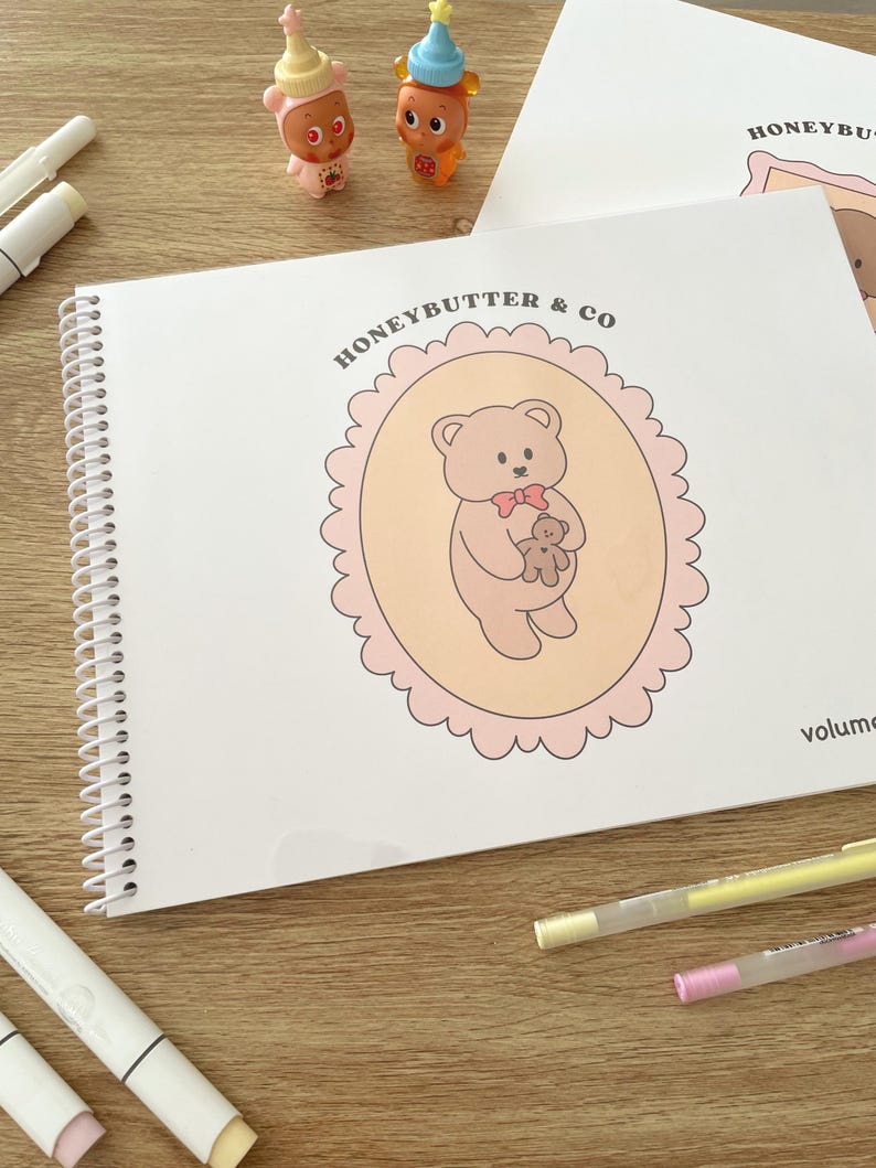 May include: A spiral-bound notebook with the text "HONEYBUTTER & CO" and a cartoon bear illustration. The bear is holding a smaller bear inside a pink scalloped oval. Two small figurines and several markers are also visible.