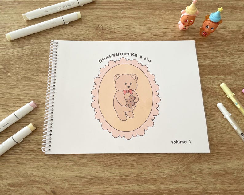 May include: A spiral-bound notebook with the text "HONEYBUTTER & CO" and a teddy bear illustration on the cover. The cover art is in a pink oval frame. The words "volume 1" are at the bottom. Markers and small figurines are scattered around the notebook.