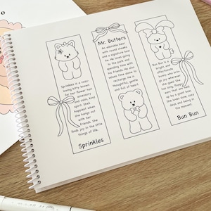 May include: A spiral-bound notebook open to a page with three illustrated bookmarks. Each bookmark features a different cartoon animal: a cat, a bear, and a bunny. Text describes each character. Markers and small figurines are also visible.