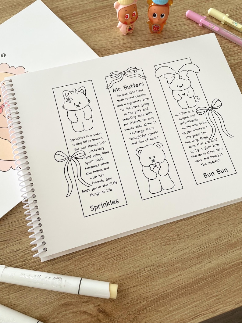 May include: A spiral-bound notebook open to a page with three illustrated bookmarks. Each bookmark features a different cartoon animal: a cat, a bear, and a bunny. Text describes each character. Markers and small figurines are also visible.