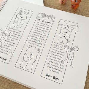 May include: A white notebook with three illustrated bookmarks featuring cartoon animals: a cat, a bear, and a bunny. Each bookmark has a description. Two small figurines are visible in the background.