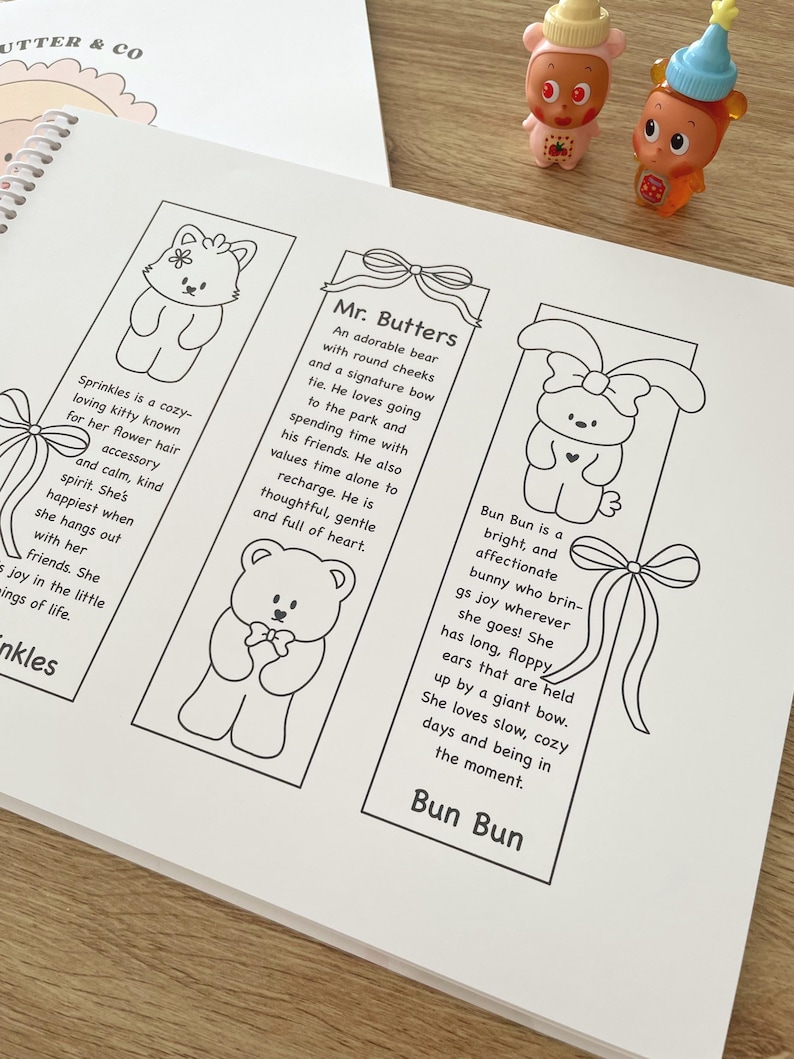 May include: A white notebook with three illustrated bookmarks featuring cartoon animals: a cat, a bear, and a bunny. Each bookmark has a description. Two small figurines are visible in the background.