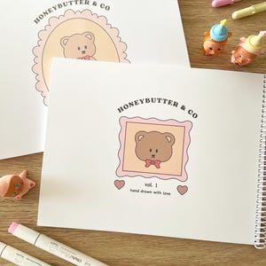 May include: White spiral-bound notebook and loose paper with a teddy bear illustration in a pink frame. The text "HONEYBUTTER & CO" and "vol. 1 hand drawn with love" are visible. Two markers and small figurines are also present.